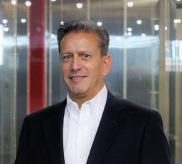 A man in a dark suit and white shirt stands indoors in front of a glass wall with lights in the background.