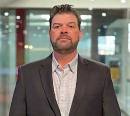 Man with short hair and beard wearing a dark blazer and light-colored shirt stands indoors in front of glass walls.